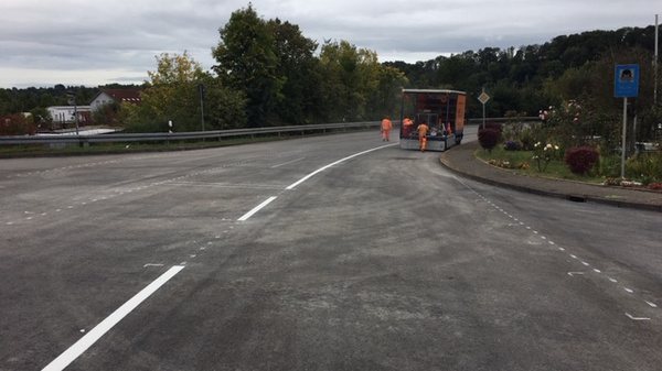Workers are seen laying down new road markings on a curved highway. The scene shows freshly painted white lines on an asphalt surface, with a truck nearby and trees lining the background, indicating road work is in progress.