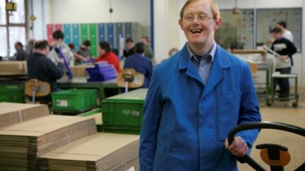 A smiling individual in a blue work jacket stands with a hand pallet truck in a warehouse setting. In the background, several people are engaged in work, sorting boxes and items in a vibrant, busy environment. The atmosphere is positive and collaborative.