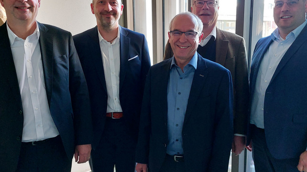 Cinq hommes portant des vêtements professionnels se tiennent debout côte à côte dans un espace intérieur lumineux. Ils sourient et semblent engagés dans une discussion. À l’arrière-plan, une plante est visible près d'une fenêtre.