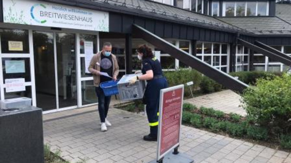 A person wearing a mask receives a container from a caregiver outside the entrance of a building labeled "Breitwiesenhaus." The setting appears to be a health or community service facility, with a pathway and greenery visible.