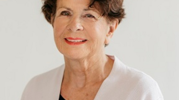A smiling older woman with curly brown hair stands against a light background. She is wearing a light-colored cardigan over a black top. Her expression is warm and friendly, conveying confidence and approachability.