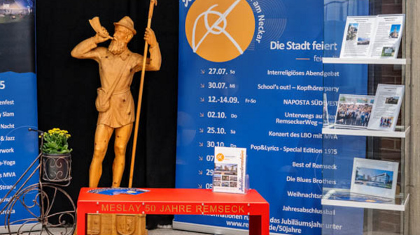 Die Abbildung zeigt eine Ausstellung mit Informationsständen, umgeben von zwei blauen Plakaten, die einen Jubiläumskalender und Ereignisse in der Stadt ankündigen. In der Mitte steht eine Statue eines Mannes, der mit einem Handzeichen auf sich aufmerksam macht, und ein roter Tisch mit weiteren Informationen und Blumen.