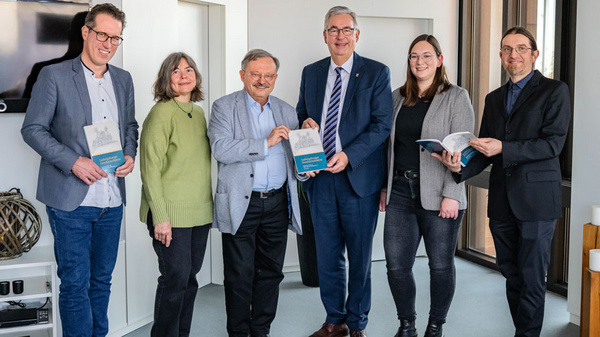Eine Gruppe von sechs Personen steht in einem modernen Raum. Sie halten Bücher in den Händen und lächeln in die Kamera. Die Szene vermittelt eine Atmosphäre der Zusammenarbeit und des Austauschs.