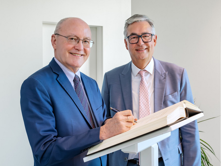 Zwei Männer stehen neben einem Schreibpult. Der links stehende Mann in einem blauen Anzug lächelt und hält einen Stift, um in ein offenes Buch zu schreiben. Der rechte Mann trägt einen grauen Anzug und sieht lächelnd in die Kamera.
