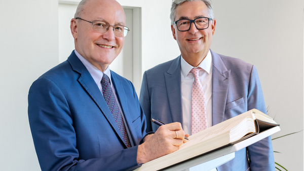 Zwei Männer stehen neben einem Schreibpult. Der links stehende Mann in einem blauen Anzug lächelt und hält einen Stift, um in ein offenes Buch zu schreiben. Der rechte Mann trägt einen grauen Anzug und sieht lächelnd in die Kamera.