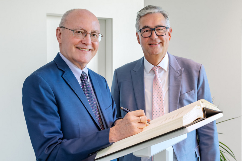 Zwei Männer stehen neben einem Schreibpult. Der links stehende Mann in einem blauen Anzug lächelt und hält einen Stift, um in ein offenes Buch zu schreiben. Der rechte Mann trägt einen grauen Anzug und sieht lächelnd in die Kamera.