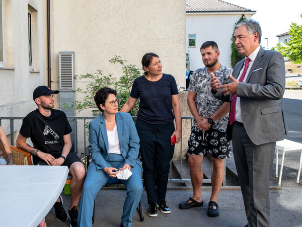 08.08.2022 - Besuch Flüchtlingsunterkunft im alten Krankenhaus Marbach mit Ministerin Marion Gentges (mitte) Eine Gruppe von fünf Personen sitzt und steht an einem Tisch im Freien. Ein Mann spricht, während die anderen ihm aufmerksam zuhören. Die Anwesenden wirken interessiert und engaged in das Gespräch. Sie sind in lässiger Kleidung und es ist ein sonniger Tag.