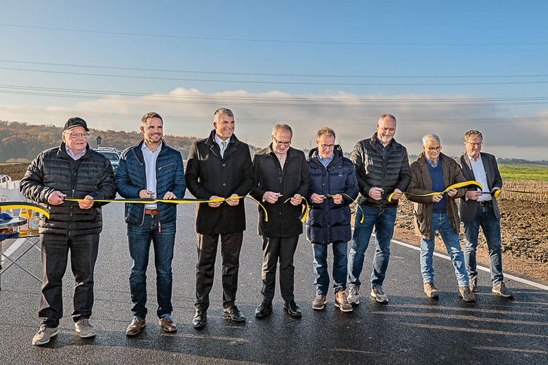 Eine Gruppe von acht Personen steht auf einer neu asphaltierten Straße und hält ein gelbes Band. Sie scheinen an einer Eröffnungszeremonie teilzunehmen. Im Hintergrund sind Felder und ein klarer Himmel zu sehen.