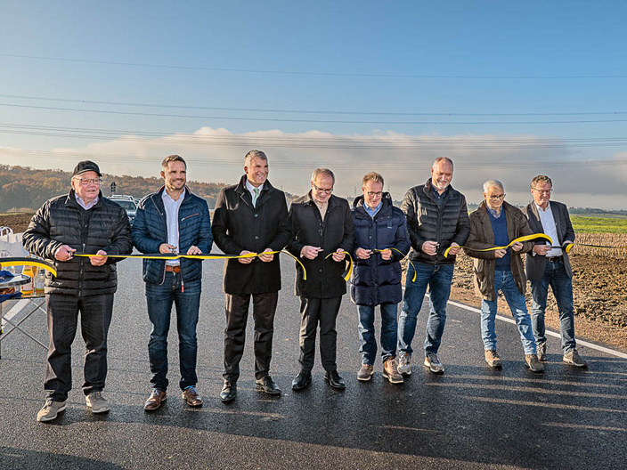 Eine Gruppe von acht Personen steht auf einer neu asphaltierten Straße und hält ein gelbes Band. Sie scheinen an einer Eröffnungszeremonie teilzunehmen. Im Hintergrund sind Felder und ein klarer Himmel zu sehen.