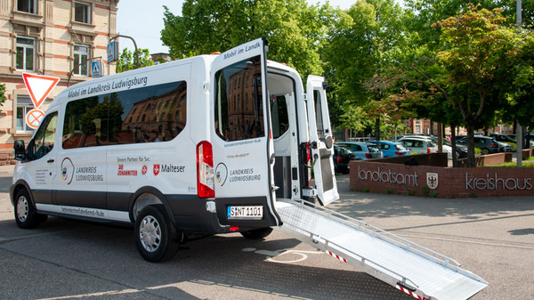 Ein barrierefreier Transporter steht im Freien. Die Rücktüren sind geöffnet und eine Rampe ist heruntergeklappt, um den Zugang zu erleichtern. Auf der Seite des Fahrzeugs ist der Schriftzug "Landkreis Ludwigsburg" zu sehen, umgeben von grünen Bäumen. (Text KI generiert)