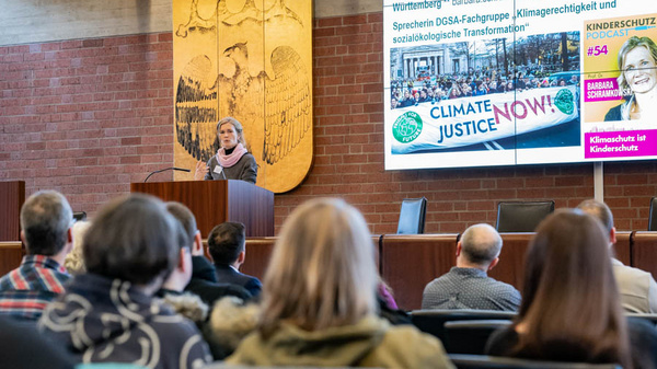 Eine Frau steht an einem Rednerpult und spricht vor einem Publikum in einem großen Saal. Im Hintergrund sind Präsentationsfolien mit dem Titel "Klimagerechtigkeit jetzt!" und Informationen zur ökologischen Transformation zu sehen. Manche Zuhörer wenden sich der Rednerin zu.