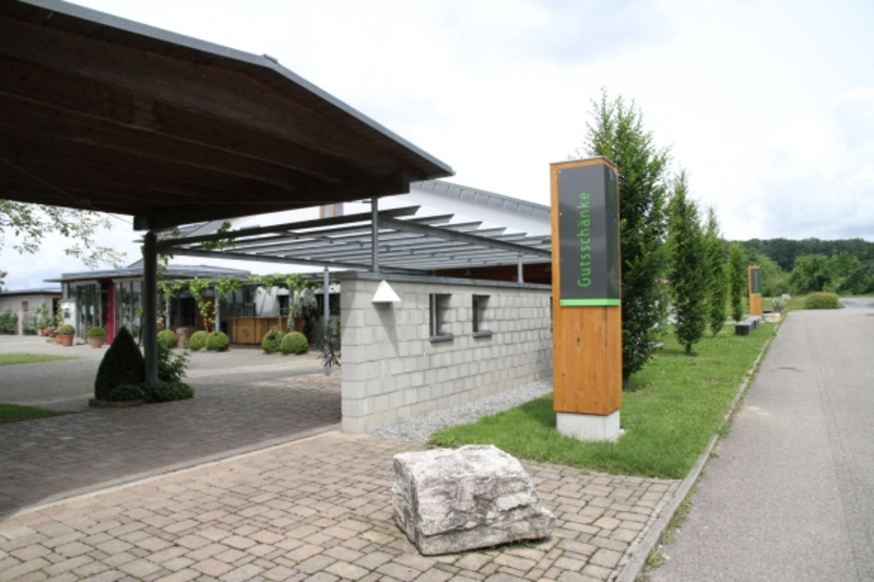 Eingang zu einem modernen Gebäude mit angrenzendem Parkplatz. Im Vordergrund eine gepflasterte Fläche und ein großes Steingewächs. Links sind gepflegte Pflanzen und rechts ein Holzschild mit der Aufschrift "Gutshof". Der Himmel ist bewölkt. (Text KI generiert)