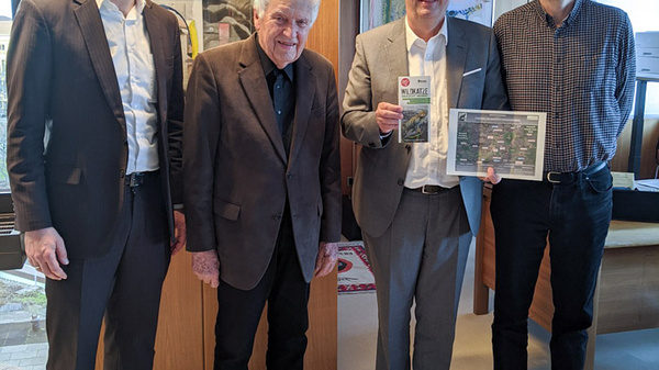 Four men stand together in an office setting. They are smiling and dressed in formal attire. One man holds a document, while another presents a photo or map. The background features wooden walls and some framed artwork, indicating a professional environment.