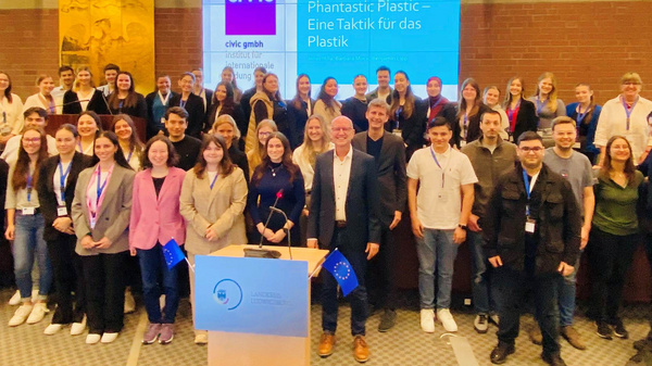 Die Abbildung zeigt eine große Gruppe von Menschen, die in einem Konferenzraum zusammenstehen, um eine Veranstaltung zum Thema Plastik zu besuchen. Im Hintergrund ist ein Bildschirm mit dem Titel "Fantastisch Plastik - Eine Taktik für das Plastik" zu sehen. Das Bild vermittelt eine Atmosphäre des Austauschs und des Engagements.