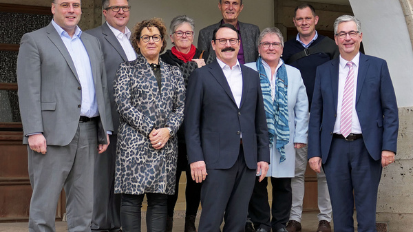 A group of nine people stands on the steps of a building under an arch. They are dressed in business casual attire, smiling and facing the camera. The setting appears to be a formal gathering or meeting.