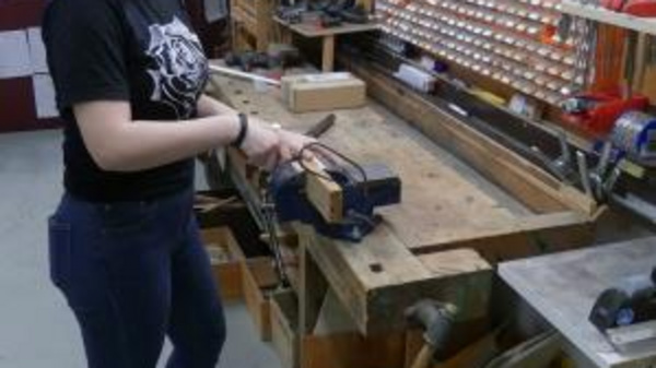 A young woman stands in a workshop, smiling as she uses a hand tool at a wooden workbench. The background features various tools organized on the wall, showcasing a creative and productive environment.