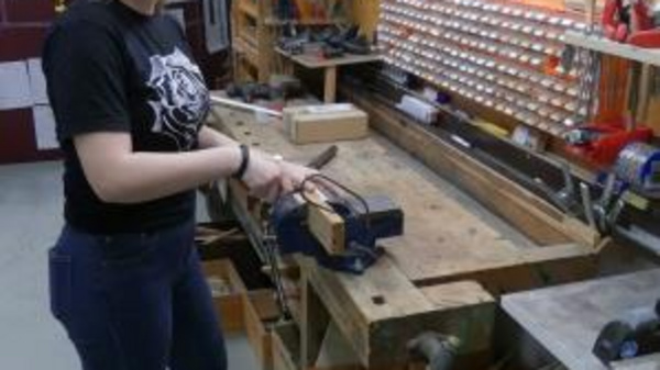 A young woman stands in a workshop, smiling as she uses a hand tool at a wooden workbench. The background features various tools organized on the wall, showcasing a creative and productive environment.