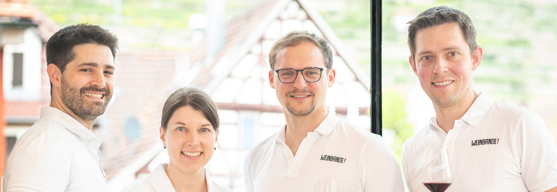 Vier Personen stehen in einem hellen Raum und halten jeweils ein Glas Wein in der Hand. Sie lächeln freundlich in die Kamera. Im Hintergrund sind Weinberge und eine helle Architektur sichtbar. Die Gruppe scheint eine gesellige Atmosphäre zu genießen.