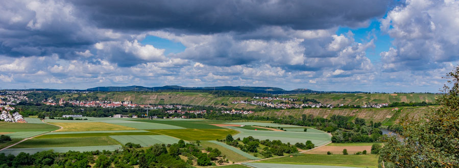 Header Bild: Eine weite Landschaft mit grünen Feldern, Weinbergen und einem Fluss im Hintergrund. Der Himmel ist von einigen Wolken bedeckt, während Teile des Himmels blau sind. Im Hintergrund sind Hügel und eine kleine Stadt sichtbar.