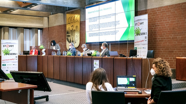 A panel of speakers, wearing masks, discusses a topic in a conference room. A presentation screen displays information, and attendees are seated at tables with laptops. Banners promoting the event are visible in the background.