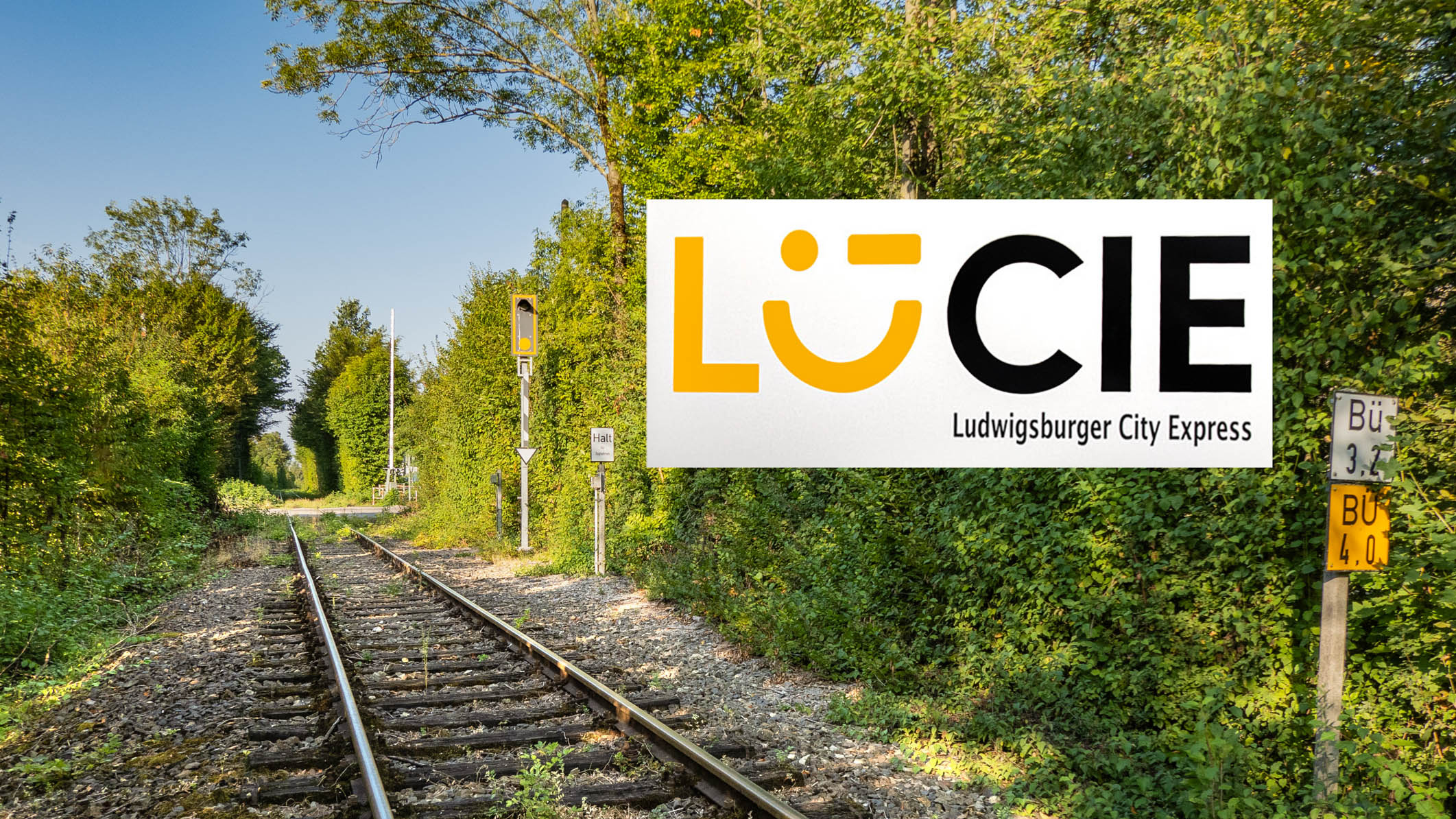 The picture shows a railroad line, to the left and right of which is greenery with trees and meadows. The Lucie light rail logo can be seen at the top right of the picture.