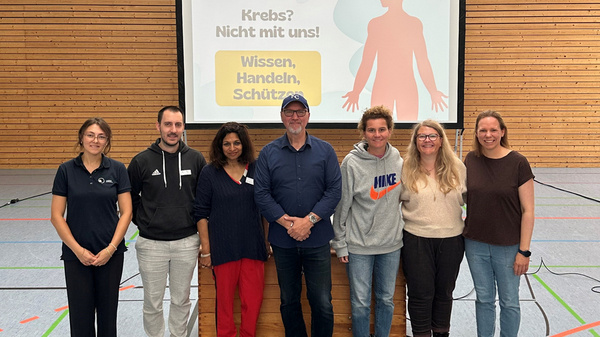 A group of seven presenters stands in front of a large screen displaying the German text "Krebs? Nicht mit uns! Wissen, Handeln, Schützen!" The setting is a gymnasium with wooden walls and a clock visible. The focus is on the theme of cancer awareness and prevention.