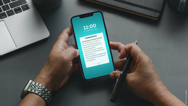 Eine Person hält ein Smartphone in der Hand, auf dem der Bildschirm eine Nachricht mit der Uhrzeit 11:00 und dem Datum 26. September 2023 zeigt. Neben der Person liegt ein Notebook und ein Kugelschreiber. (Text KI generiert)