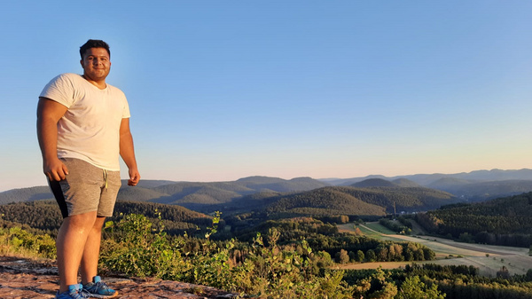 Un homme se tient sur un promontoire rocheux, regardant un vaste paysage de collines et de vallées verdoyantes sous un ciel clair. Il porte un t-shirt blanc et un short, avec des baskets bleues. L'ambiance est paisible et naturelle, évoquant une belle journée en plein air.