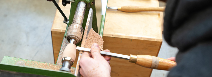 Header Bild: Ein Handwerker bearbeitet mit einem Holzschnittwerkzeug ein Werkstück an einer Drehmaschine. Der Fokus liegt auf der Hand des Handwerkers, die präzise das Werkzeug ansetzt, während Holzspäne um ihn herum auf der Maschine liegen.
