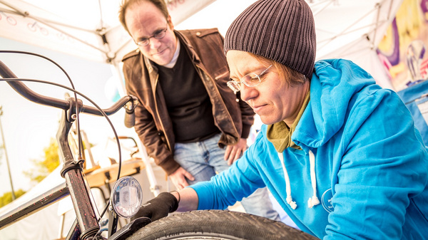 A woman wearing an aqua hoodie and a beanie is repairing a bicycle tire, focused on her task. A man in a brown jacket watches her intently, observing her work. They are outdoors, under a tent, suggesting a community or event setting related to cycling.