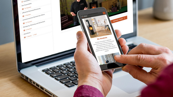 Ein Mensch hält ein Smartphone in der Hand, während er auf einen Laptop schaut. Auf dem Laptop-Bildschirm ist eine Webseite mit dem Titel „Einleitung“ sichtbar. Im Hintergrund steht eine Pflanze in einem modernen Topf. Der Arbeitsplatz ist ordentlich und minimalistisch eingerichtet.