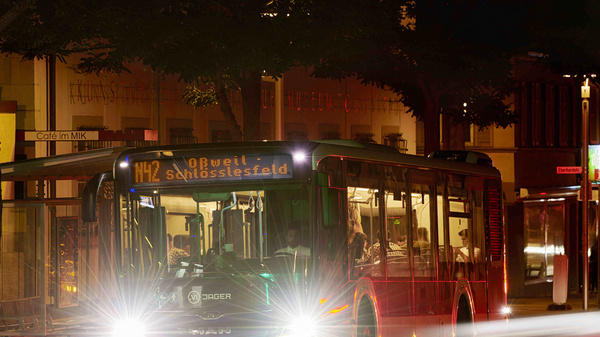Ein roter Stadtbus steht an einer Haltestelle in der Nacht. Die Frontlichter leuchten hell und die Anzeige zeigt "H2 Schwisselfeld". Im Hintergrund sind unscharfe Bäume und eine weitere Haltestelle zu erkennen.