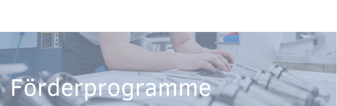 Ein Mensch arbeitet an einem Laptop, umgeben von technischen Geräten. Der Text „Förderprogramme“ ist prominent platziert und deutet auf Informationen zu finanziellen Unterstützungsmöglichkeiten hin.