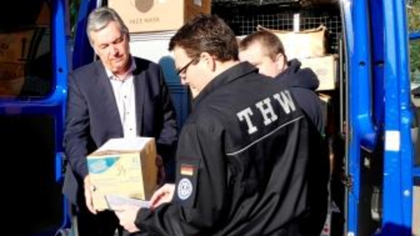 A man in a suit hands a box to a volunteer in THW attire at the back of a blue van. Several cardboard boxes are visible inside the van, indicating a relief or support effort. The scene captures a moment of teamwork and assistance.