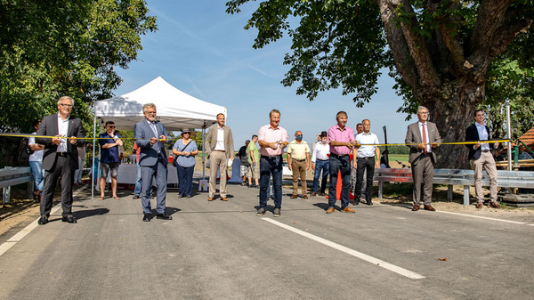 A group of people stands on a road holding a large ribbon, preparing to cut it during an event. Several individuals are gathered around, and a tent is visible in the background, indicating a formal ceremony, likely for a new road opening or similar celebration.