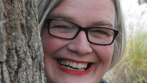 Eine lächelnde Frau mit grauem Haar und Brille versteckt sich teilweise hinter einem Baumstamm. Sie trägt ein schwarzes Oberteil und einen grünen Schal. Ihr Gesicht strahlt Freude aus, und im Hintergrund sind unscharf Pflanzen zu sehen.