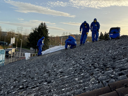 Arbeiter in blauen Overalls sind auf einem schrägen Dach beschäftigt. Sie führen Reparaturen durch und verwenden dabei verschiedene Werkzeuge. Im Hintergrund sind Bäume und ein Parkplatz sichtbar. (Text KI generiert)
