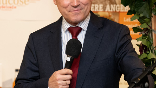 A man in a suit stands at a podium, smiling and holding a microphone. He appears to be giving a speech or presentation, engaging with the audience. The background contains banners with text related to food products.