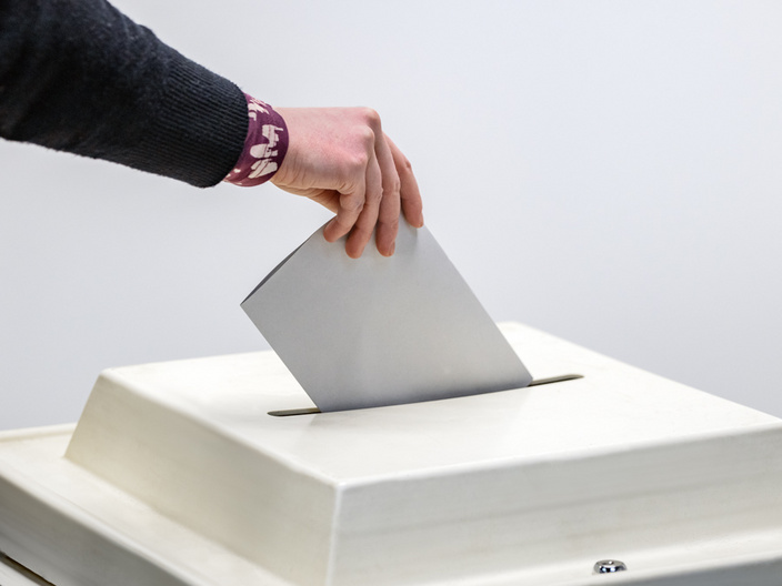 Eine Hand wirft einen Stimmzettel in eine Wahlurne. Der Stimmzettel ist grau und die Urne hat eine quadratische Öffnung. Die Szene symbolisiert die Teilnahme an einer Wahl.