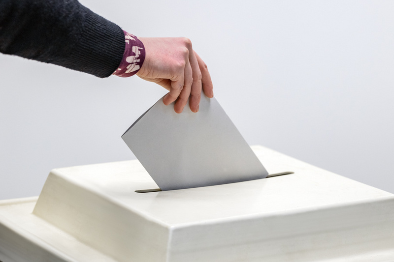 Eine Hand wirft einen Stimmzettel in eine Wahlurne. Der Stimmzettel ist grau und die Urne hat eine quadratische Öffnung. Die Szene symbolisiert die Teilnahme an einer Wahl.