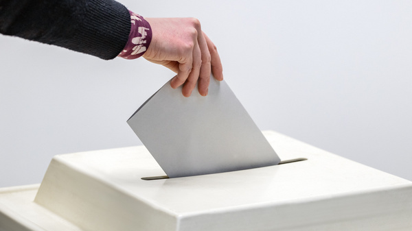 Eine Hand wirft einen Stimmzettel in eine Wahlurne. Der Stimmzettel ist grau und die Urne hat eine quadratische Öffnung. Die Szene symbolisiert die Teilnahme an einer Wahl.