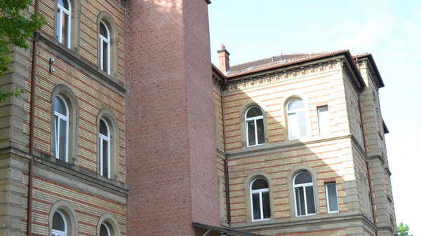 Immagine di un edificio storico a tre piani, con una facciata in mattoni e grandi finestre ad arco. La struttura presenta dettagli architettonici distintivi e si trova in un contesto verdeggiante, con alberi nelle vicinanze. Il cielo è parzialmente coperto da nuvole.