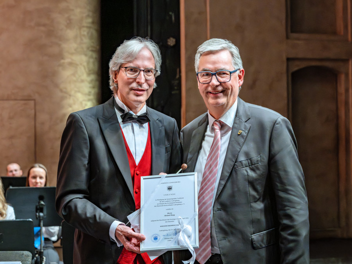 Zwei Männer stehen auf einer Bühne. Der Mann links trägt einen Frack mit roter Weste und hält ein Zertifikat in der Hand. Der Mann rechts trägt einen Anzug und lächelt. Im Hintergrund sind verschwommen weitere Menschen zu sehen, die auf einer Aufführung sitzen.