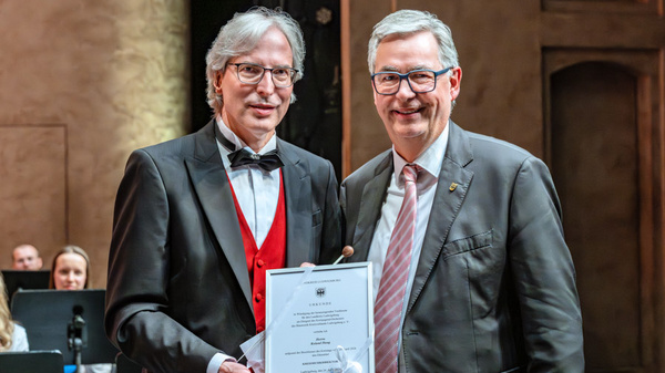 Zwei Männer stehen auf einer Bühne. Der Mann links trägt einen Frack mit roter Weste und hält ein Zertifikat in der Hand. Der Mann rechts trägt einen Anzug und lächelt. Im Hintergrund sind verschwommen weitere Menschen zu sehen, die auf einer Aufführung sitzen.