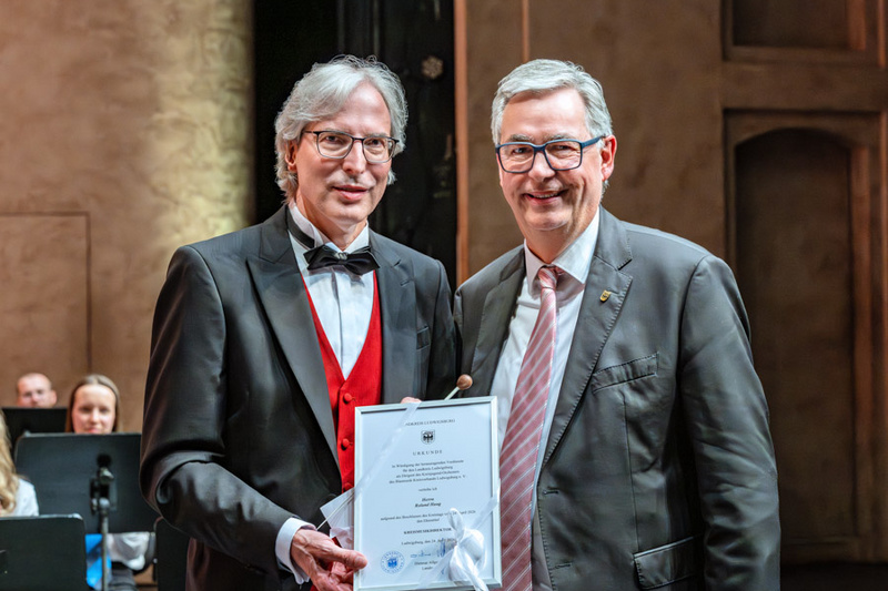 Zwei Männer stehen auf einer Bühne. Der Mann links trägt einen Frack mit roter Weste und hält ein Zertifikat in der Hand. Der Mann rechts trägt einen Anzug und lächelt. Im Hintergrund sind verschwommen weitere Menschen zu sehen, die auf einer Aufführung sitzen.