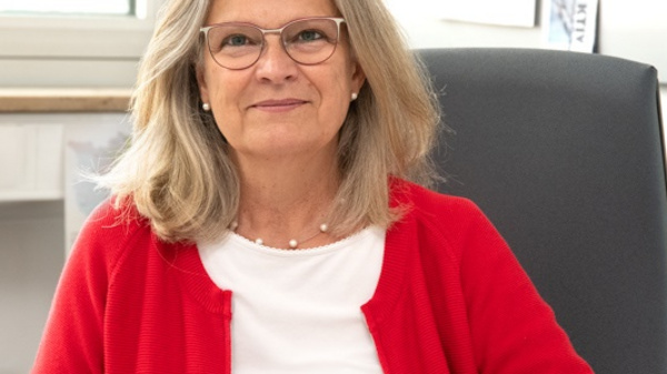 A woman with long, wavy hair and glasses smiles while seated at a desk. She wears a red cardigan over a white shirt. The office background includes papers and a visible window, creating a bright and professional atmosphere.