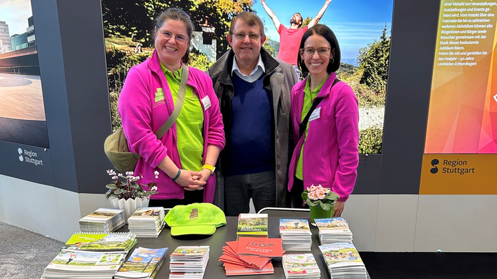 Eine Gruppe von drei Personen steht an einem Stand mit der Aufschrift "rundum aktiv region stuttgart". Die beiden Frauen tragen leuchtend pinke Jacken, während der Mann in der Mitte lässig gekleidet ist. Auf dem Tisch sind verschiedene Broschüren und ein grünes Hut-Accessoire sichtbar.