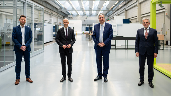 Vier Männer stehen in einem modernen Industrieumfeld. Sie tragen formelle Kleidung und lächeln in die Kamera. Im Hintergrund sind Maschinen und Arbeitsbereiche zu erkennen. Die Männer scheinen an einer Veranstaltung oder Besprechung teilzunehmen.