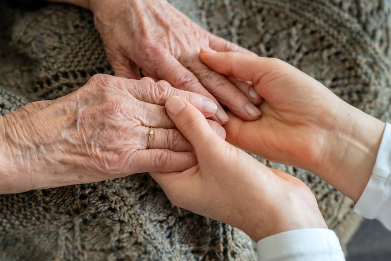 Zwei Hände halten sich, eine ältere Hand mit sichtbaren Adern und einer goldenen Ring, während eine jüngere Hand zart darüber greift. Die Geste symbolisiert Zuneigung und Unterstützung zwischen den Generationen.
