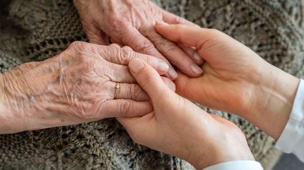 Zwei Hände halten sich, eine ältere Hand mit sichtbaren Adern und einer goldenen Ring, während eine jüngere Hand zart darüber greift. Die Geste symbolisiert Zuneigung und Unterstützung zwischen den Generationen.